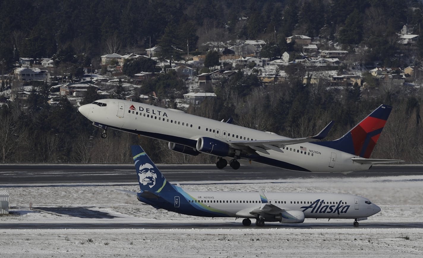 A Delta Air Lines plane takes off above a taxiing Alaska Airlines plane, Tuesday, Feb. 5, 2019, on a snow-bordered runway at Seattle-Tacoma International Airport in Seattle.