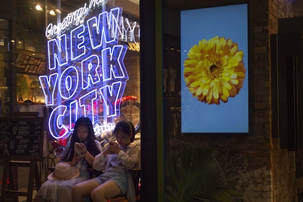 In this Thursday, July 5, 2018 photo, girls sit in front of an American cosmetics brand's shop window display reading "Greetings from New York City" a