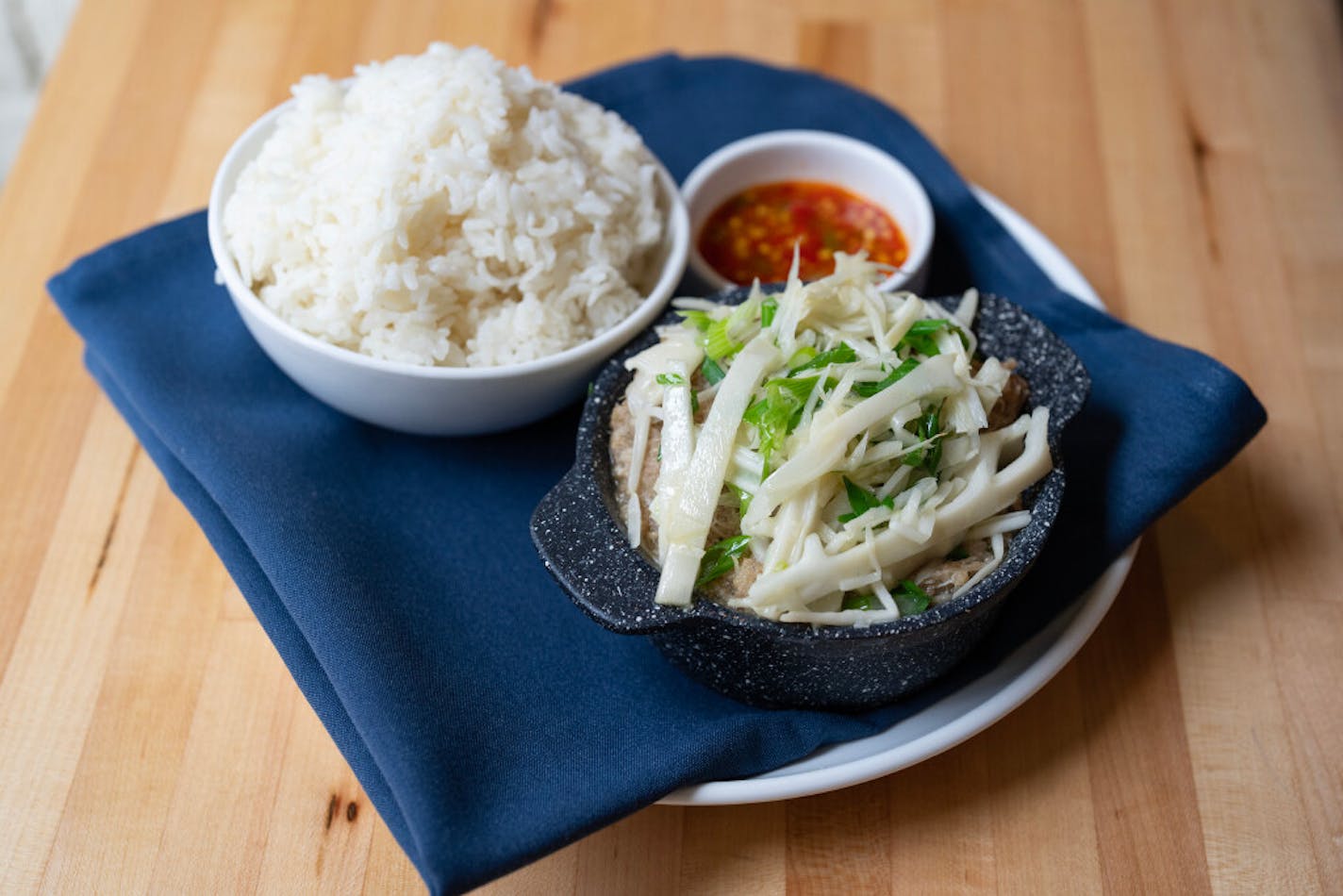 A cast iron dish mounded ith pork, garnishe with long strips of ginger and green onions served alongside a bowl of rice and a small container of vibrant chili sauce.