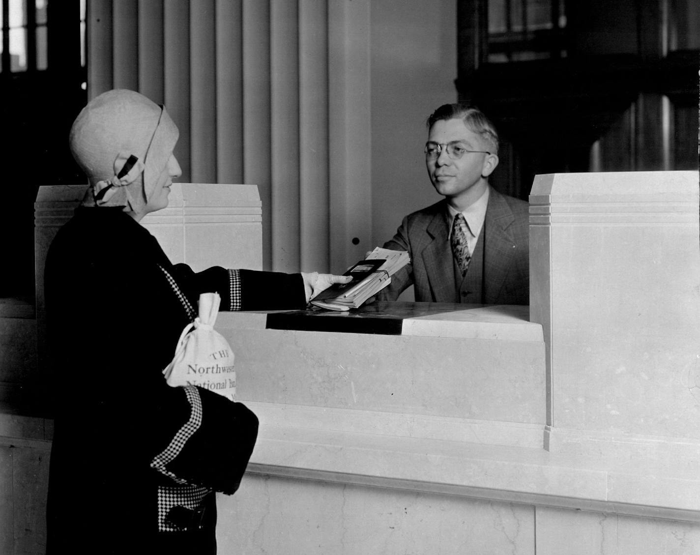 Mildred Mullowney, 3132 Columbus_Avenue, is the first customer to make a deposit at Northwestern National Bank after its removal to the new building. A. A. Dahlberg is the teller. This photograph shows a typical teller's station. They are all open counters, no cages or bars, tellers stationed back of marble counter with cash in money desk with pull-down locking cover. There are 115 tellers' stations in all. Commercials Photographs