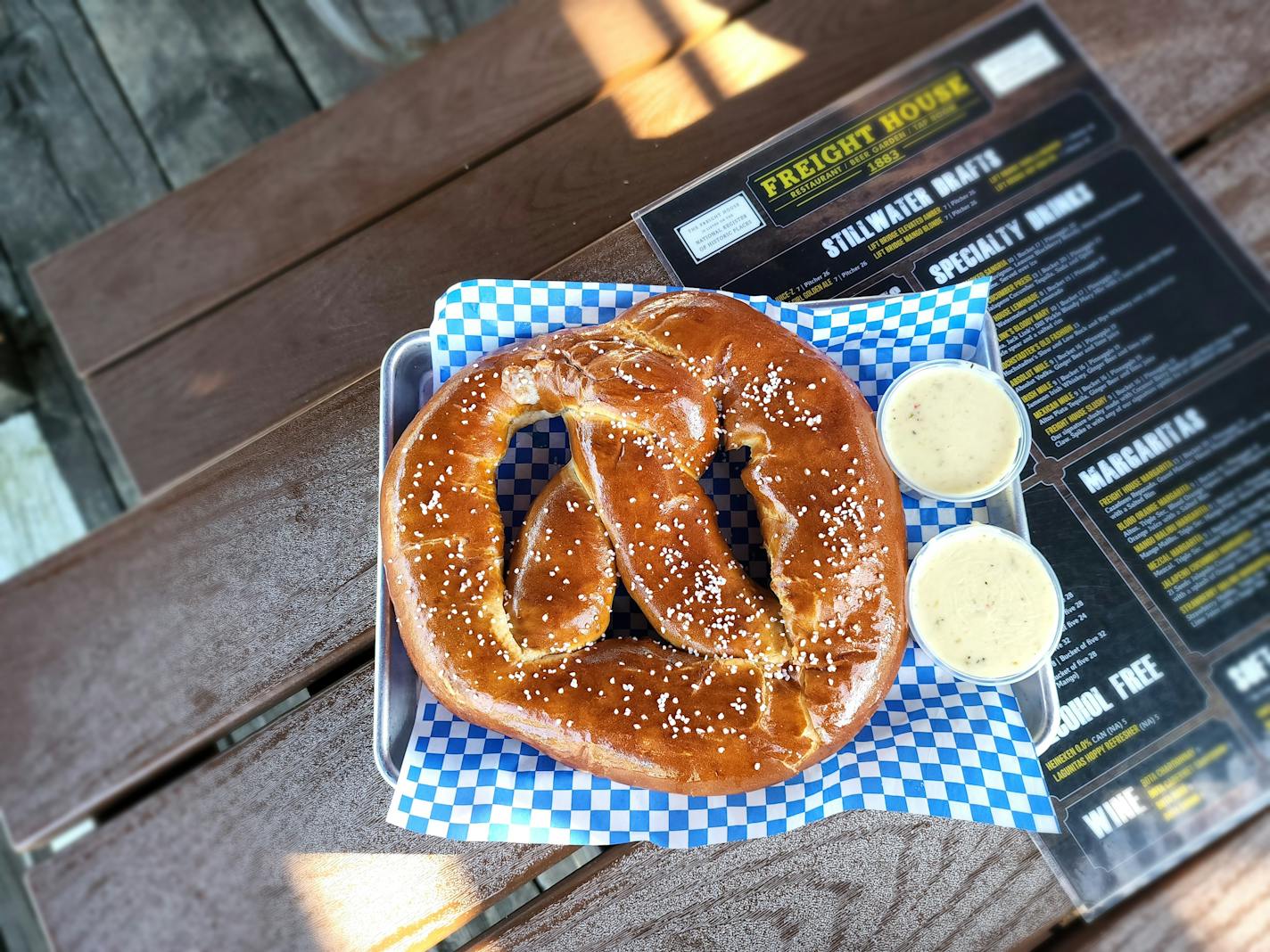 A large salted pretzel on a blue and white checkered paper with two containers of cheese sauce, on a wooden picnic table showing a Freight House menu