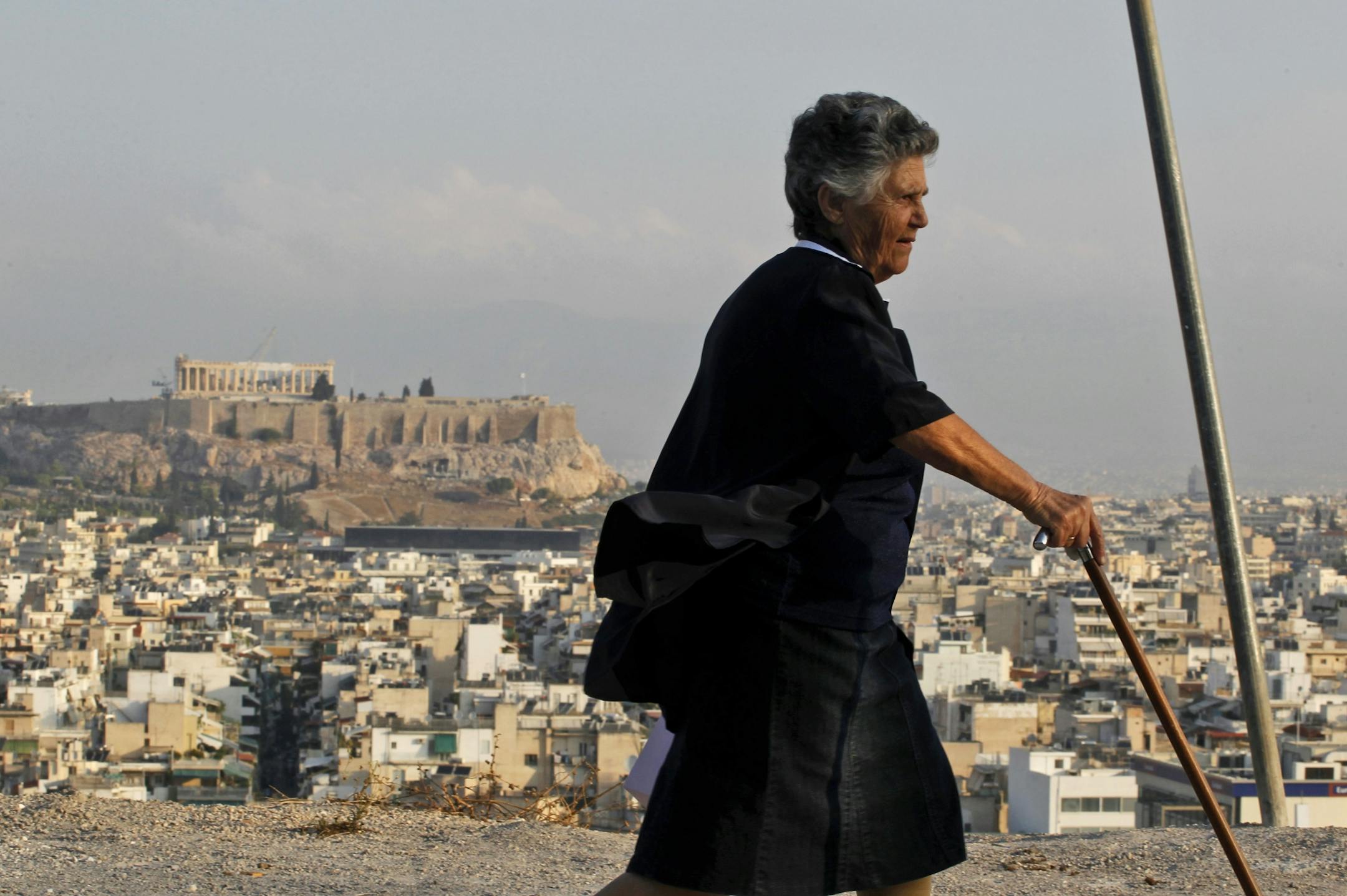An elderly woman walks by the ancient Acropolis hill and the city of Athens.