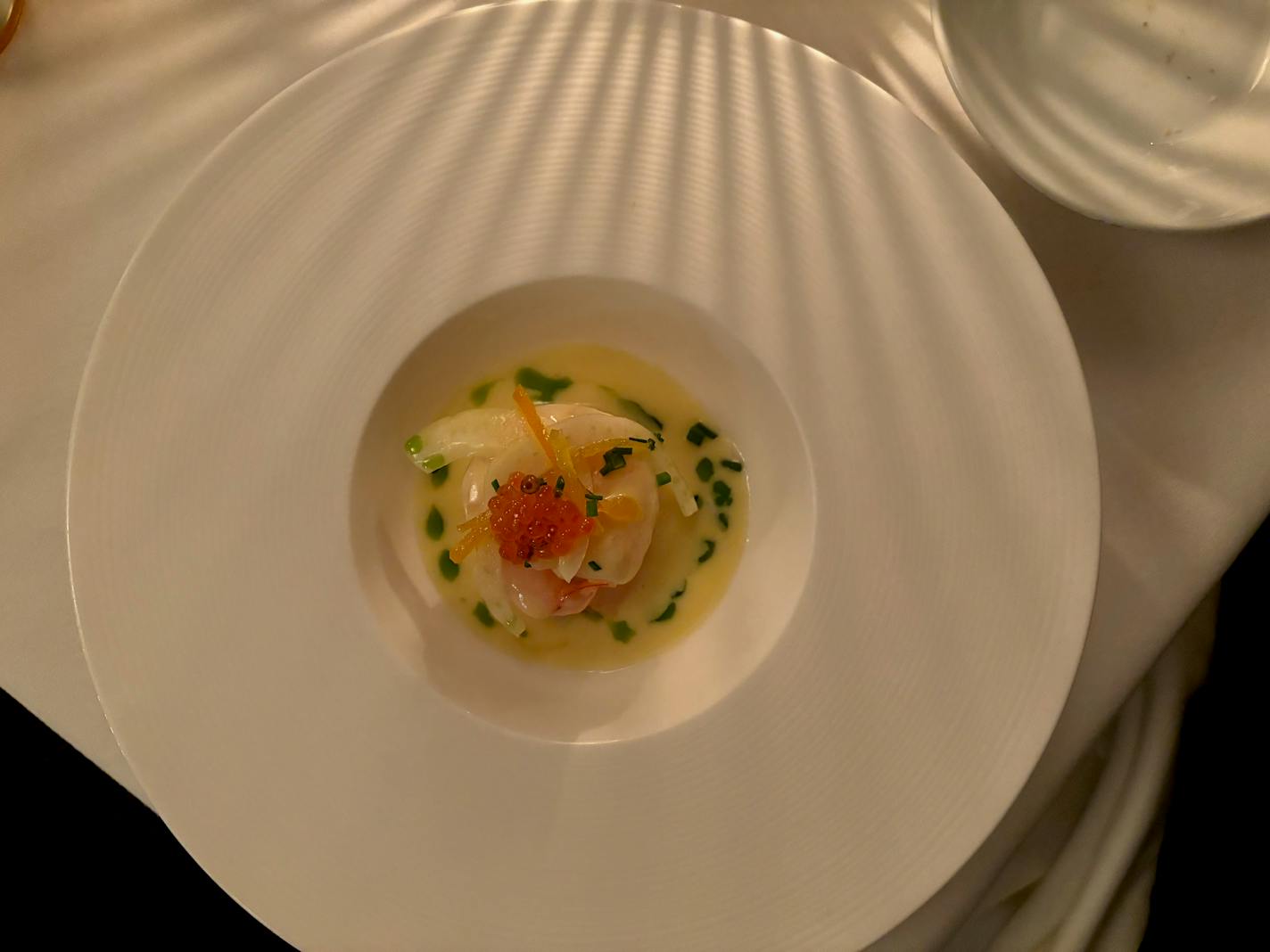 In a dimly lit restaurant a large white plate has a small depression in the middle in which a poached shrimp lays in a shallow pool of cream and topped with slivers of orange and garnished with a sprinkling of chives.