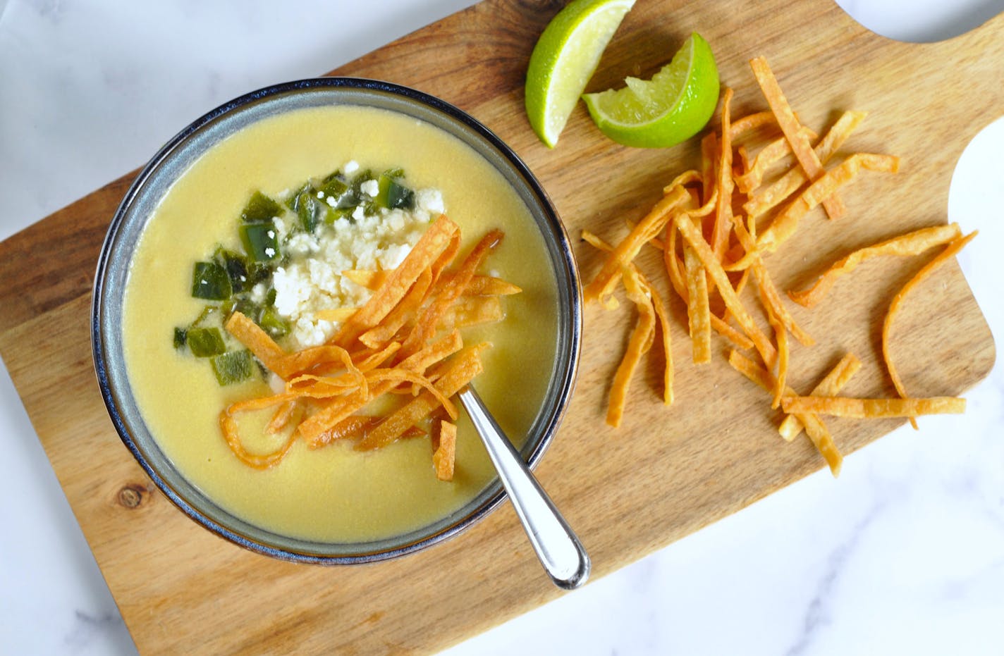 A bowl of soup on a cutting board. The bowl is filled with golden corn soup topped with poblanos, queso fresco, crispy tortilla strips with a squeeze of lime.