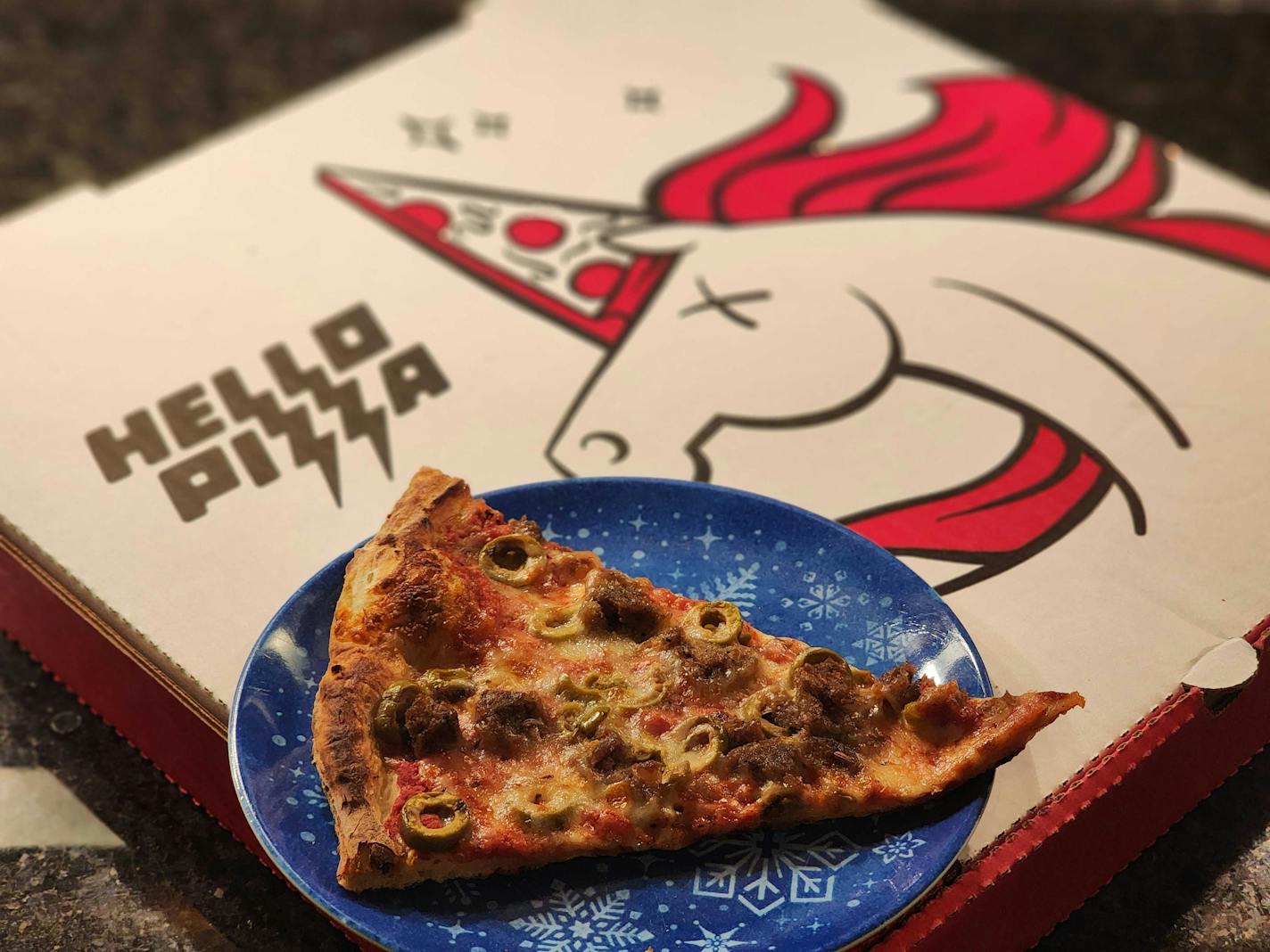 A pink and white pizza box with an image of a unicorn, and a blue plate on top of it with a slice of sausage and olive pizza