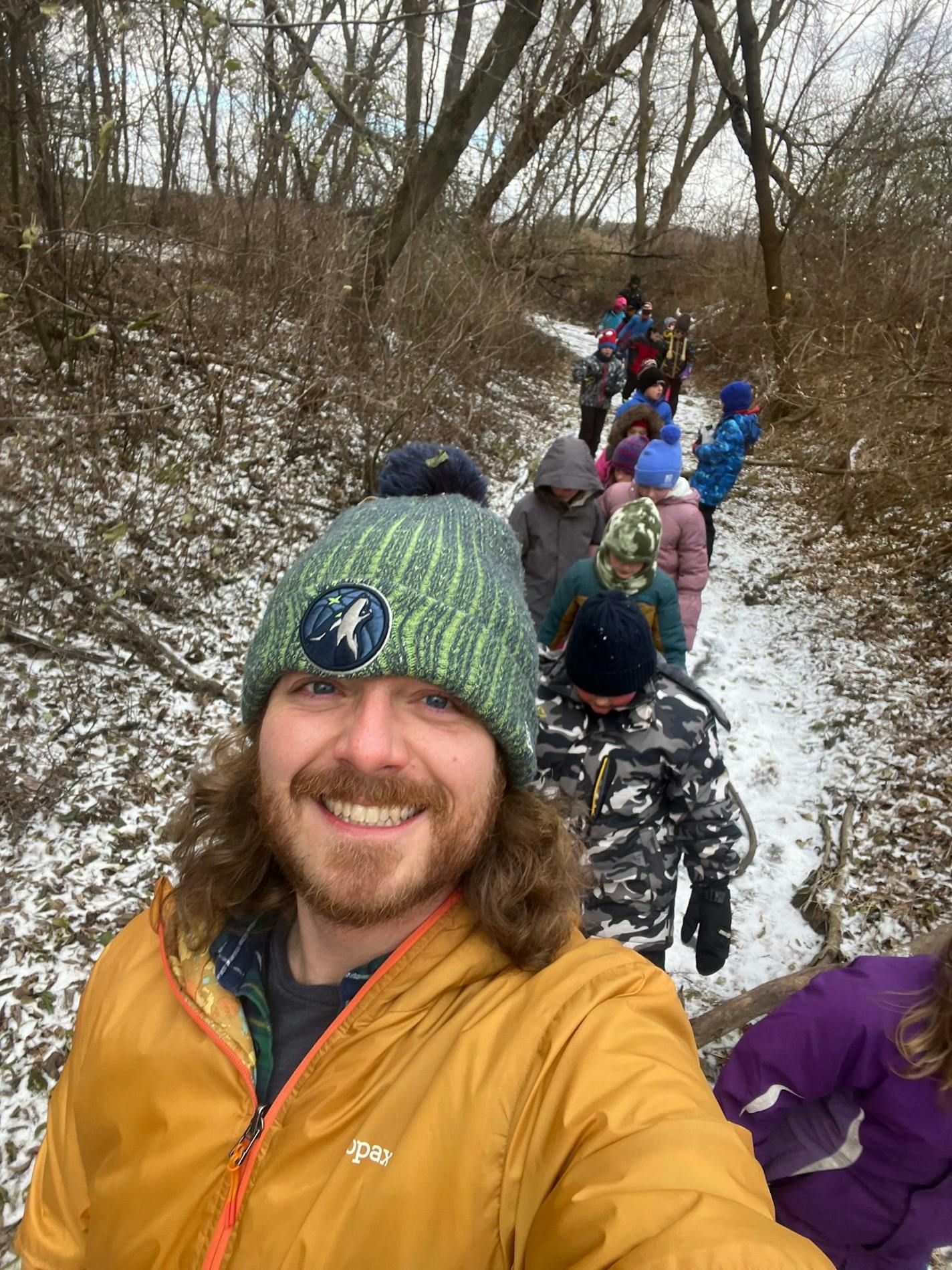 A 30-something bearded man wearing a jacket and stocking cap smiles for a selfie on a snow-covered trail surrounded by bare trees. A class of bundled-up kids in second and third grade follows him.