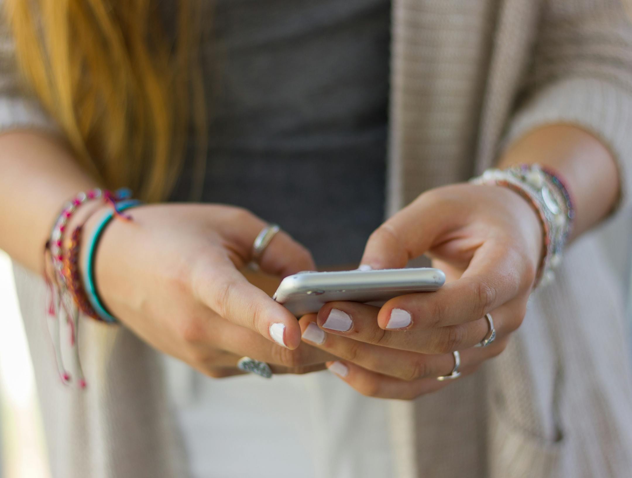 Cell phone on hands of teenage blonde girl with withe painted nails, bracelets and rings. Millennial generation, social media, smartphone addiction concept