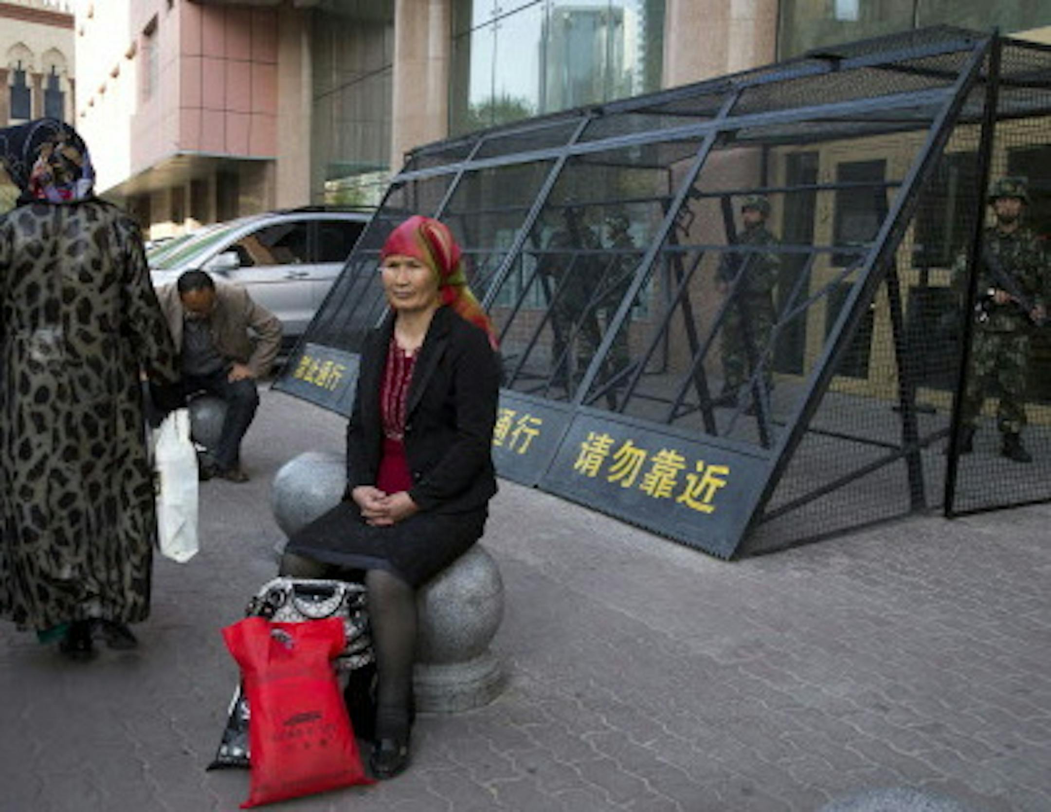 FILE - In this May 1, 2014 file photo, a Uighur woman rests near a cage protecting heavily armed Chinese paramilitary policemen on duty in Urumqi in China's northwestern region of Xinjiang. While most Chinese can easily obtain passports, eroding barriers to travel have thrown into relief a new pattern showing that entire ethnic groups deemed potentially risky to the leadership - such as Muslim Uighurs and Buddhist Tibetans - are largely being barred. By denying them opportunities for jobs, educa
