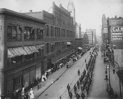 A parade on 4th Street passing by the Grand Arcade in 1907. The towered building to the rear is the old St. Paul City Hall-Ramsey County Courthouse, razed in 1933.