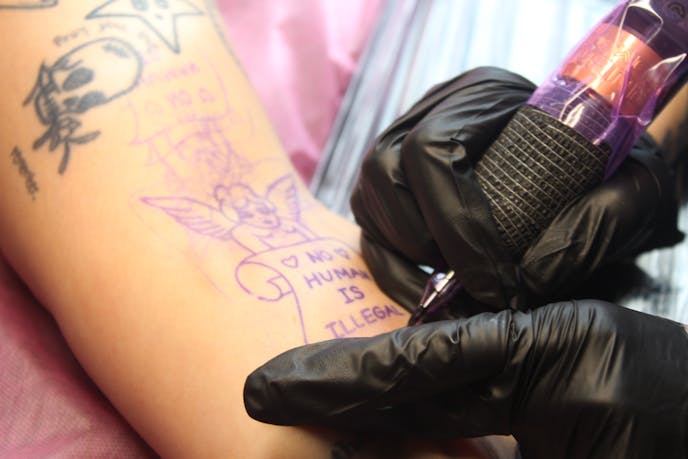 A customer at Kaleidoscope Tattoo Collective waits as an artist inks a design reading "No Human is Illegal" into the customer's arm.