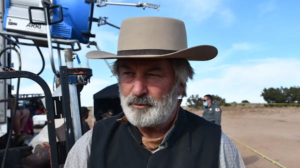 Alec Baldwin wearing a cowboy hat and old-timey Western clothes outdoors with a big camera and other film equipment on his left.