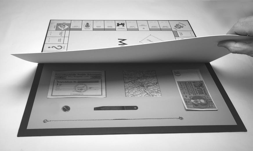 Black and white image shows a Monopoly board game, with the cover lifted to reveal money and other items hidden under it.