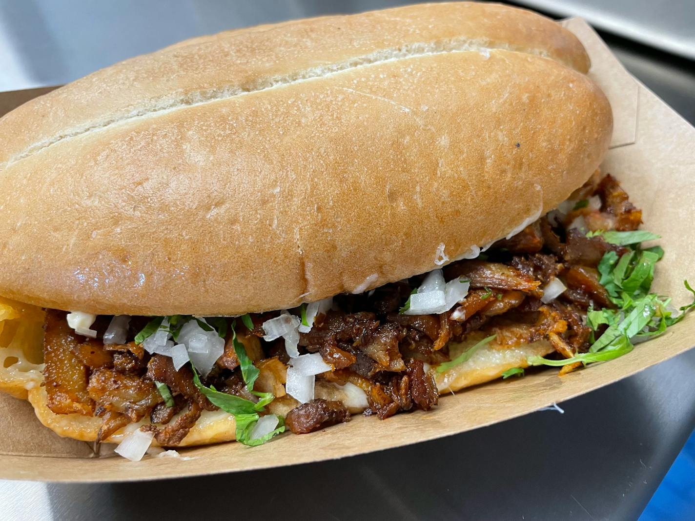 A close up photo of the torta, with a split roll of crusty bread stuffed with al pastor meat. Onions and cilantro are spilling out the front of it.