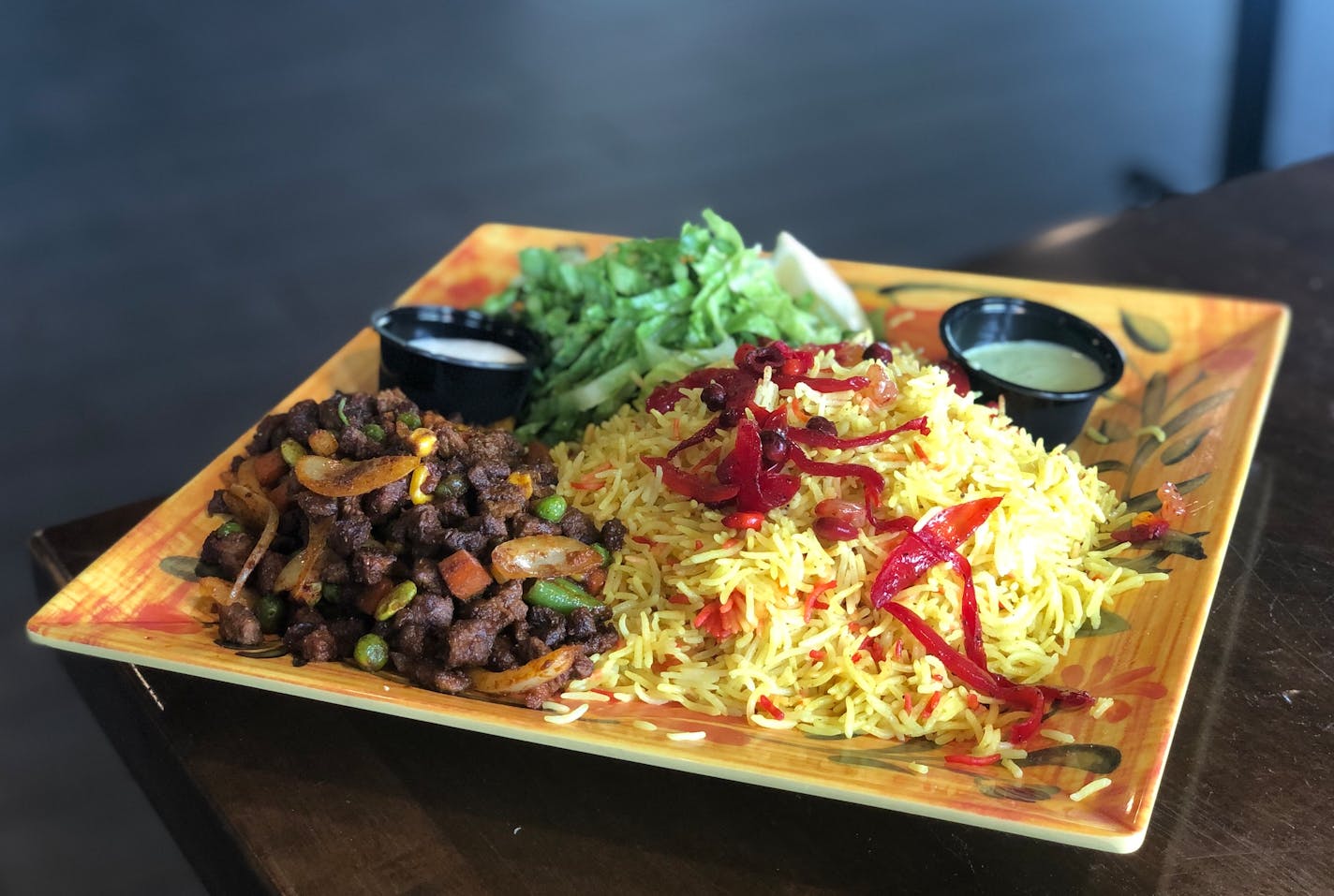 A colorful yellow square plate with beef suqaar — cubed beef with sautéed vegetables — and Somali rice and lettuce.