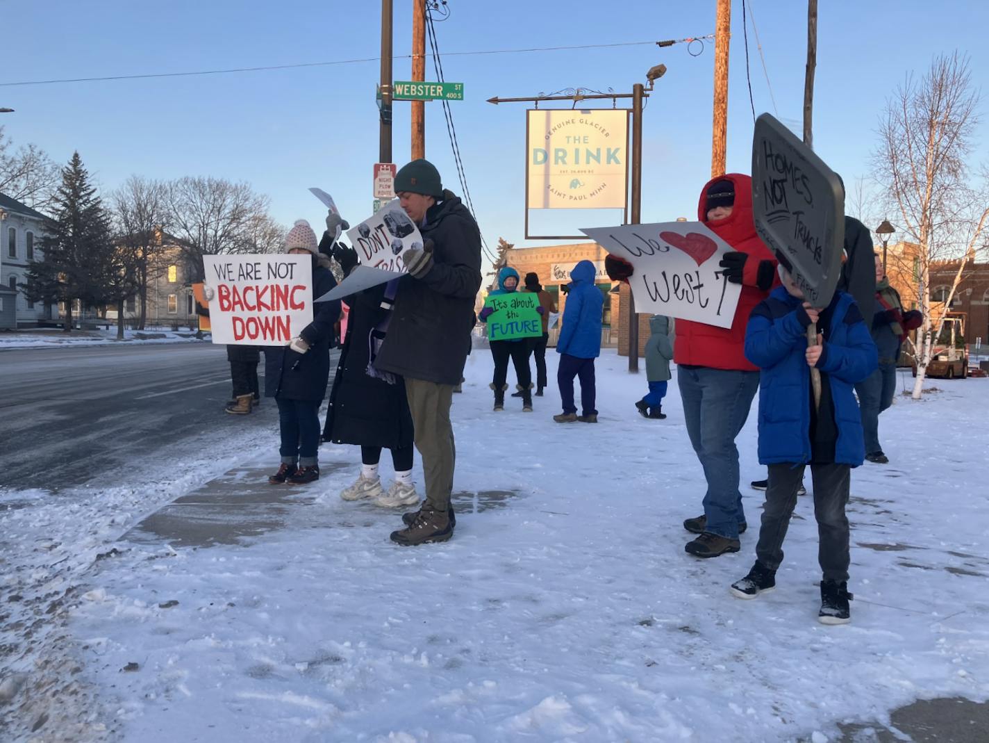 A small group of people in winter coats and hats, standing on a snow-covered with signs reading "We are not backing down," "Homes not Trucks" and "We [heart] West 7th."