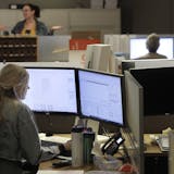 Architectural designer Erica Shannon, front, works at a computer as accounting manager Andrea Clark, top, speaks with a colleague at the design firm Bergmeyer, Wednesday, July 29, 2020, at the company's offices, in Boston. Around the U.S,. office workers sent home when the coronavirus took hold in March are returning to the world of cubicles and conference rooms and facing certain adjustments: masks, staggered shifts, limits on how many people can be there at any one time, spaced-apart desks, da
