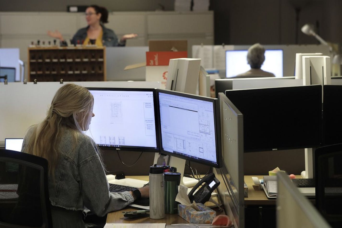 Architectural designer Erica Shannon, front, works at a computer as accounting manager Andrea Clark, top, speaks with a colleague at the design firm Bergmeyer, Wednesday, July 29, 2020, at the company's offices, in Boston. Around the U.S,. office workers sent home when the coronavirus took hold in March are returning to the world of cubicles and conference rooms and facing certain adjustments: masks, staggered shifts, limits on how many people can be there at any one time, spaced-apart desks, da