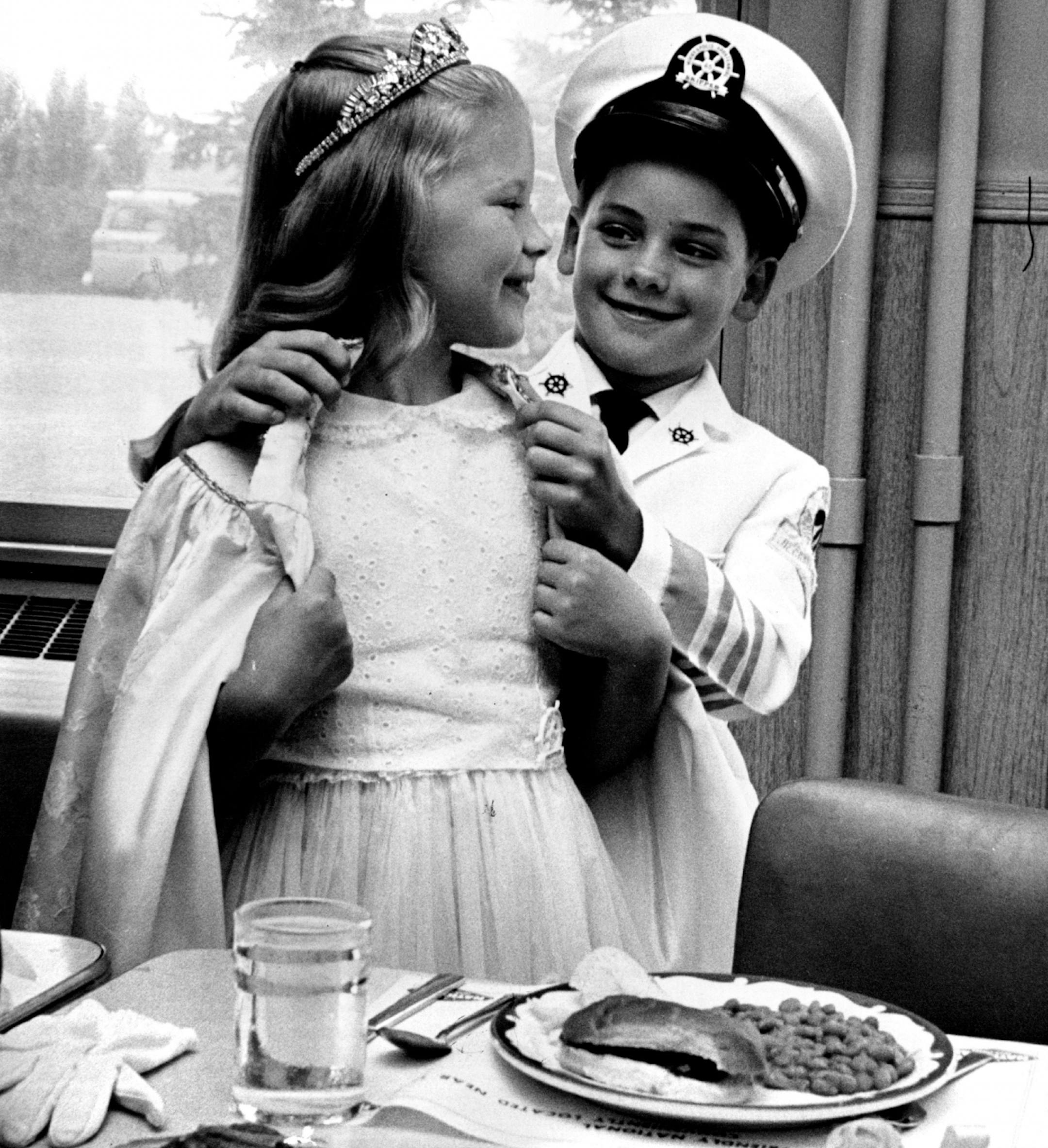 July 23, 1965: Junior Royalty 'Puts on the Dog' -- Junior Commodore Michael Audbercht, 8, 6131 Dupont Av. S., assisted his royal junior Queen, 7-year-old Linda Johnson, 3985 NE. Hayes St., Columbia Heights, with her robe Thursday, below, as they prepared to sit down to hot dogs and potato chips at the Aquantennial Junior Queen and commodore Luncheon at National Tea Co.