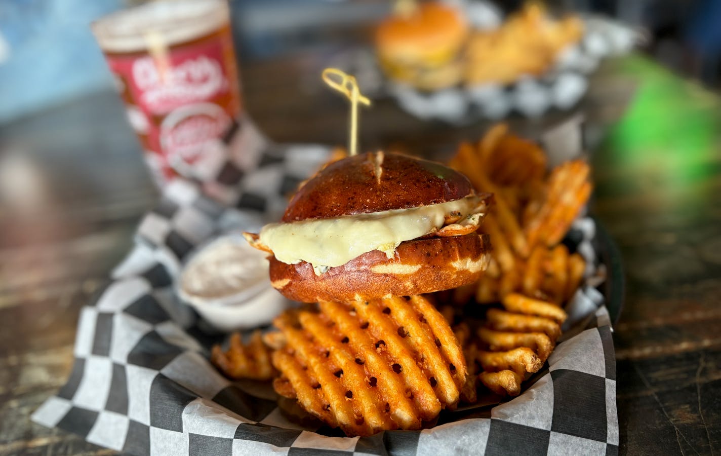 Grilled chicken sandwich with spinach artichoke dip and cheese on a pretzel bun served with waffle fries and seasoned sour cream in a basket on a sunny table top with a light beer in the background.
