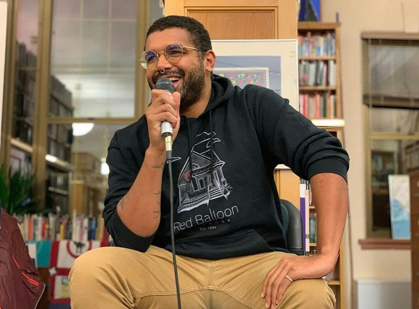 author Ty Chapman is pictured at a book event, seated and holding a microphone while wearing a shirt from the Red Balloon book store