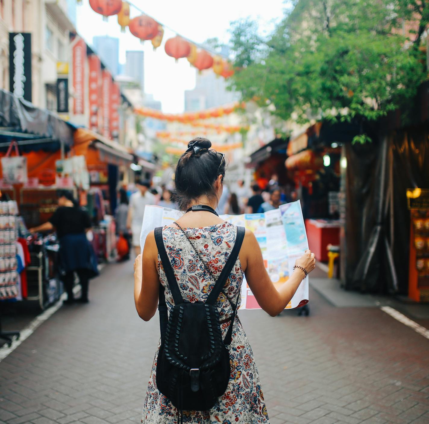 Rear view image of a young brunette woman. She is enjoying the walk and exploring the city, wearing a casual but fashionable dress, sightseeing and shopping on the Singapore street market. She is holding a large city map, checking out where to go next. ORG XMIT: 680563449